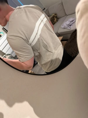 Beige t-shirt med vita ränder - Snygg beige t-shirt med två vita horisontella ränder över bröstet. Klassisk rund halsringning och korta ärmar. Perfekt för en clean och stilren look. Tillverkad i mjuk bomull för skön komfort.