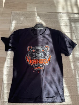 Svart Kenzo t-shirt med tigertryck - Svart t-shirt från Kenzo med ikoniskt tigertryck i orange, grått och vitt på bröstet. Klassisk rund halsringning och korta ärmar. Perfekt för dig som vill sticka ut med en streetstyle-look och gillar exklusiva märken.