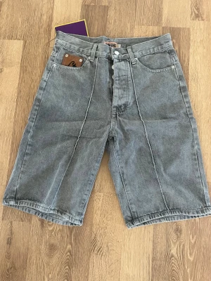 Grå denimshorts från Corteiz - Snygga grå denimshorts från Corteiz med klassisk femficksdesign och knäppning framtill. Shortsen har en loose passform och är tillverkade i slitstark bomull. Perfekta för en avslappnad streetstil.