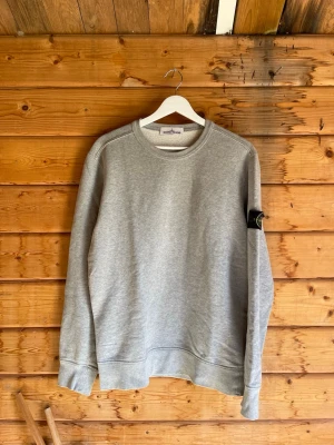 Grå sweatshirt från Stone Island - Säljer en stilren grå sweatshirt från Stone Island med klassisk rund halsringning och ribbade muddar. Tröjan har den ikoniska svarta logotypen på vänster ärm. Perfekt för en avslappnad och trendig look.