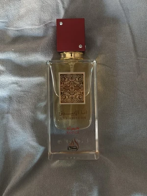 Lattafa Ana Abiyedh Rouge parfym - Säljer en elegant parfym från Lattafa, Ana Abiyedh Rouge. Flaskan är genomskinlig med gulddetaljer och har en fyrkantig röd kork dekorerad med små stenar. Designen är lyxig med arabiska mönster och text på framsidan. Perfekt för dig som gillar exklusiva dofter. Pris kan diskuteras