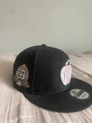 Svart Rosa New Era Yankees keps - Säljer en svart snapback keps från New Era med New York Yankees logga i vitt och rosa äpple framtill. Kepsen har platt skärm, broderade detaljer på sidan och klassisk 9FIFTY modell. Perfekt för dig som gillar streetwear och sportig stil.
