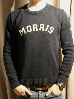 Tunn Marinblå stickad tröja Morris - Snygg marinblå stickad tröja från Morris med vit broderad logga på bröstet. Tröjan har rund halsringning och ribbade muddar vid ärmslut och nederkant. Perfekt för dig som gillar klassisk och stilren design.