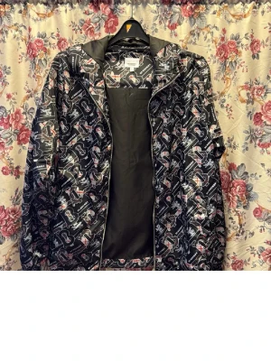 Svart vindjacka med Burberry-mönster - Snygg svart vindjacka från Burberry med ikoniskt monogrammönster i vitt och beige. Jackan har huva, dragkedja framtill och elastiska muddar vid ärmarna. Tillverkad i lätt och glansigt material som passar perfekt för blåsiga dagar.