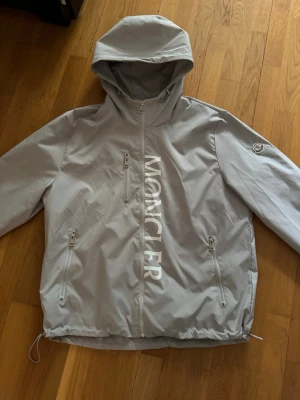 Ljusgrå vindjacka från Moncler - Snygg ljusgrå vindjacka från Moncler med huva och dragkedja framtill. Jackan har stora vita Moncler-tryck på bröstet, två sidofickor med dragkedja och justerbar nederkant. Perfekt för blåsiga dagar och streetwear-stil.