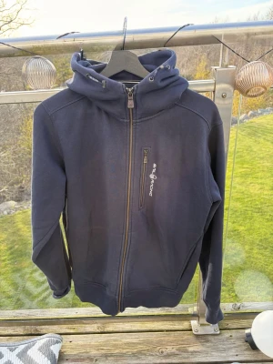 Mörkblå hoodie från Sail Racing - Mörkblå hoodie från Sail Racing med dragkedja framtill och en bröstficka med logga. Hoodien har en stor huva med dragsko och broderad detalj bak. Tillverkad i mjukt och bekvämt material, perfekt för chill dagar. Aldrig använd och är i mycket fint skick, skriv vid minsta fundering🙌🏻