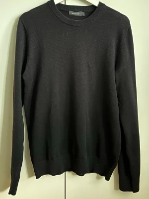 Svart tröja från Dressmann - En stilren svart tröja från Dressmann med rund halsringning och långa ärmar. Tröjan är enkel och klassisk i modellen, perfekt för lager-på-lager och har ribbade muddar vid ärmslut och nederkant. Materialet är mjukt och bekvämt.