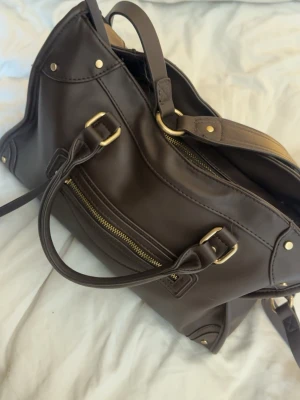 Mörkbrun handväska med gulddetaljer - Snygg mörkbrun handväska i slätt läder med guldfärgade metalldetaljer och dragkedja framtill. Väskan har dubbla handtag och axelrem, samt dekorativa nitar i hörnen.