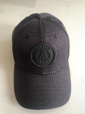 Svart PSG keps med logga - Snygg svart keps från Paris Saint-Germain med upphöjd PSG-logga framtill och diskret mönster med texten 'PARIS' över hela kepsen. Justerbar baktill och tillverkad i bomullsmix. Perfekt för dig som gillar fotboll och streetstyle.