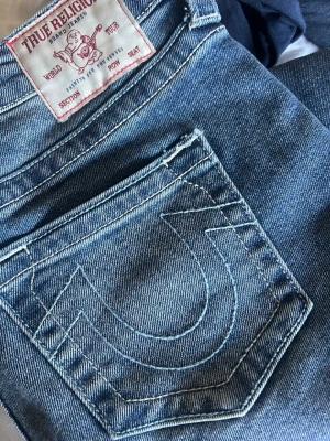 True Religion ljusblå jeans - Säljer ett par jättesnygga  ljusblå jeans från True Religion med deras klassiska broderade bakficka och låg midja. Jeansen har utsvängda ben och är tillverkade i denim med snygga kontrastsömmar. Perfekta för alla tillfällen!