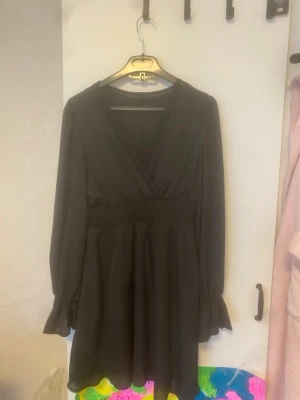 Svart kort omlottklänning - Svart kort klänning med omlott-skärning och v-ringning. Långa ärmar med lätt volang vid ärmslut och markerad midja. Perfekt för dig som gillar en stilren och enkel look.
