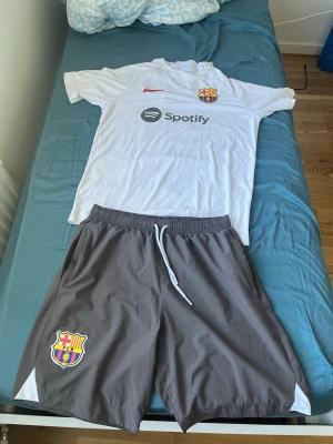 FC Barcelona träningsset Nike - Säljer ett FC Barcelona träningsset med vit t-shirt och grå shorts. T-shirten har klubbmärke, Nike-logga och Spotify-tryck framtill. Shortsen är grå med klubbmärke och vit dragsko. Perfekt för fotbollsträning eller gym. Materialet är lätt och andas.
