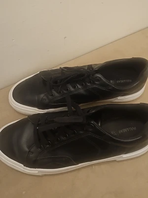 Svarta sneakers från Pull&Bear - Svarta sneakers från Pull&Bear med klassisk låg profil och vita sulor. Skorna har svarta skosnören, diskret design och är tillverkade i syntetmaterial med läderlook. Perfekt för dig som gillar stilrena och enkla sneakers.