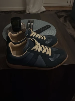 Mörkblå sneakers från Maison Margiela - Snygga mörkblå sneakers från Maison Margiela med beige detaljer och klassiska gum-yttersulor. Skorna har beige snörning och en låg profil som ger en clean och stilren look. Materialmix av läder och mocka för extra premiumkänsla. Använda några gånger men har inga skavanker. Otorligt bekväma. Priset kan sänkas vid snabb affär