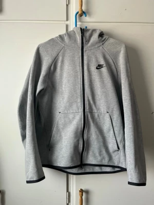 Grå zip hoodie från Nike - Snygg grå hoodie från Nike med dragkedja framtill och svart Nike-logga på bröstet. Hoodien har huva, två stora fickor och svarta detaljer vid dragkedja och ärmslut. Tillverkad i mjukt och bekvämt material, perfekt för en avslappnad stil.