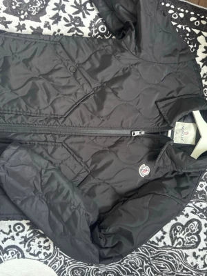 Svart dunjacka från Moncler - Snygg svart dunjacka från Moncler med quiltat mönster och dragkedja framtill. Jackan har två snedställda fickor med tryckknapp och klassisk Moncler-logga på bröstet. Perfekt för kalla vinterdagar.