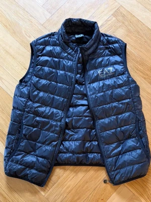 Svart dunväst från EA7 Emporio Armani - Snygg svart dunväst från EA7 Emporio Armani med quiltad design och hög krage. Västen har dragkedja framtill och EA7-logga på bröstet. Perfekt för lager-på-lager och streetstyle.