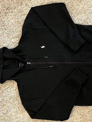 Svart hoodie från Polo Ralph Lauren - Snygg svart hoodie från Polo Ralph Lauren med dragkedja framtill och klassisk vit logga på bröstet. Hoodien har huva med dragsko och ribbade muddar vid ärmar och nederkant. Tillverkad i mjukt bomullsmaterial för en skön känsla. skicket är 9/10 använd fåtal ggr. 