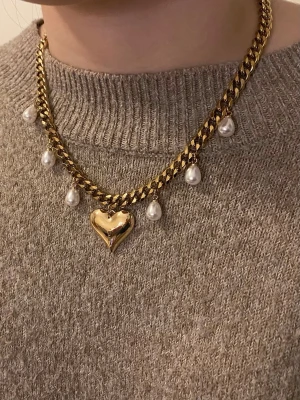 Guldigt halsband med pärlor och hjärta - Säljer ett chunky guldigt halsband med hängande vita pärlor och ett stort hjärtformat hänge i mitten. Kedjan är bred och har en cool, modern look som verkligen sticker ut. Perfekt för dig som gillar statement-smycken och vill ha något unikt.
