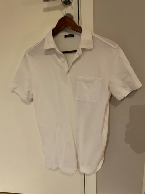 Vit piketröja Massimo Dutti - Klassisk vit pikétröja från Massimo Dutti med korta ärmar och bröstficka. Piketröjan har en stilren krage och knappar framtill. Perfekt för en clean och enkel look. Tröjan är nästintill oanvänd.