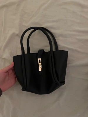 Svart handväska med guldfärgat spänne - Stilren svart handväska med två handtag och ett elegant guldfärgat spänne framtill. Väskan har en klassisk och enkel design, perfekt för att bära dina viktigaste saker. Materialet är slätt och väskan har en strukturerad form.