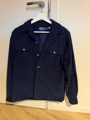 Ralph Lauren manchester overshirt  - Stilren marinblå overshirt i manchestertyg från Polo by Ralph Lauren. Klassisk krage, knäppning framtill och två bröstfickor med lock och knapp. Perfekt lager-på-lager-plagg med avslappnad passform och snygg struktur. Knappt använd.