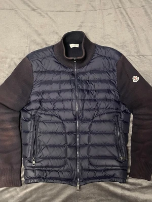Svart pufferjacka från Moncler - Snygg svart pufferjacka från Moncler med quiltad front och stickade ärmar. Jackan har hög krage, två dragkedjefickor och Moncler-logga på vänster ärm. Perfekt för kalla vinterdagar och ger en stilren look.