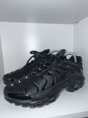 Nike Air Max Plus Triple Black - Svarta Nike Air Max Plus sneakers med mesh- och syntetöverdel, svarta snören och distinkta vågformade detaljer. Sulan har synliga Air Max-enheter och Tn-logga undertill. Snygg och sportig modell med chunky look och helsvart färgsättning.