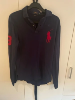 Mörkblå långärmad skjorta Polo Ralph Lauren - Mörkblå långärmad skjorta från Polo Ralph Lauren med röd logga på bröstet och röd siffra 3 på ärmen. Klassisk krage och knappar framtill. Perfekt för en sportig och stilren look.