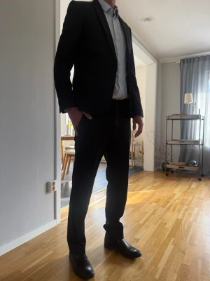 Svart kostym med kavaj och byxor - Stilren svart (antracit) kostym med klassisk kavaj och matchande byxor. Kavajen har smal passform, spetsiga slag och knäppning framtill. Perfekt för dig som vill ha en enkel och tidlös look. Skjorta ingår ej. Märke: bläck