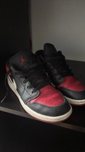 Air Jordan 1 Low Svart/Röd - Säljer ett par Air Jordan 1 Low sneakers i svart och rött läder med vit swoosh och röd yttersula. Klassisk siluett med svarta skosnören och perforerad tåbox. Jumpman-logga på plösen. Perfekta för dig som gillar streetwear och ikoniska sneakers.