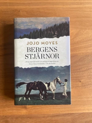 Bergens stjärnor av Jojo Moyes - Säljer romanen 'Bergens stjärnor' av Jojo Moyes. Boken har ett hårt omslag med ett naturmotiv i blå, grön och beige ton samt illustration av hästar och en kvinna. Handlingen utspelar sig i 1930-talets Kentucky och handlar om kvinnlig vänskap och mod.