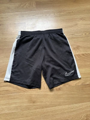 Svarta Nike shorts med vita sidor - Svarta sportiga shorts från Nike med vita paneler längs sidorna och klassisk Nike-logga framtill. Elastisk midja för skön passform. Perfekta för träning eller chill. Tillverkade i lätt och luftig polyester.