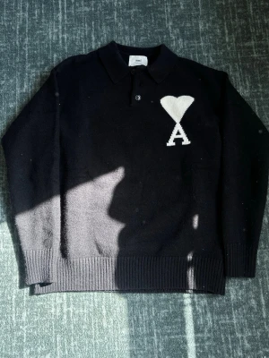 Svart stickad tröja från Ami - Svart stickad tröja från Ami med klassisk krage och två knappar framtill. På bröstet finns ett vitt hjärta och bokstaven A. Tröjan har ribbstickade muddar och är perfekt för en stilren look. Passar M/L