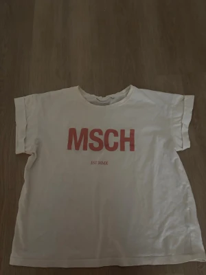 Vit t-shirt med röd MSCH-logga - Vit t-shirt från MSCH med röd tryckt logga på bröstet och texten EST MMX under. T-shirten har korta ärmar och en avslappnad passform. Perfekt för en chill och stilren look.