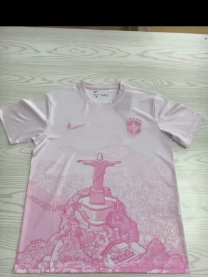 Nike Brasil rosa t-shirt med tryck - Snygg rosa t-shirt från Nike med Brasil-logga och ikoniskt tryck av Kristusstatyn i Rio de Janeiro. T-shirten har rund hals, korta ärmar och är tillverkad i ett lätt och sportigt material. Perfekt för dig som gillar unik design och sportig stil.