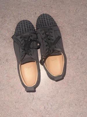 Svarta sneakers med nitar från Louboutin - Säljer ett par gråa sneakers från Christian Louboutin med coola gråa nitar på tån och klassisk röd sula. Skorna har gråa skosnören och ovandel i textil. Insidan är beige och sulan har den ikoniska Louboutin-signaturen.