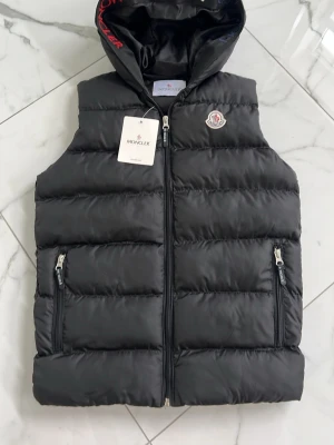 Svart dunväst från Moncler Barn - Snygg svart dunväst från Moncler med huva och dragkedja framtill. Västen har två fickor med dragkedja och Moncler-logga på bröstet. Tillverkad i glansigt material och fylld med dun för extra värme. Perfekt för lager på lager och streetstyle.