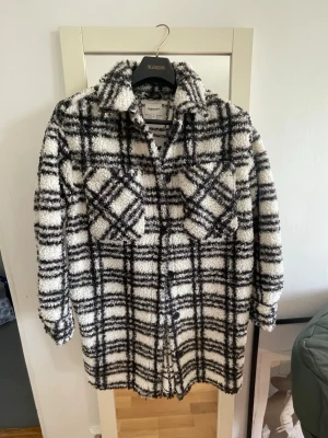 Vår -Kapa  rutmönstrad overshirt - Säljer en fluffig overshirt från Kari Traa i svartvitt rutmönster. Jackan har två stora bröstfickor med lock, knäppning framtill och en oversized passform. Materialet är mjukt och teddy-liknande, perfekt för lager på lager under kyliga dagar.