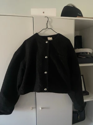 Svart cardiganjacka med pärlknappar - En svart, croppad cardiganjacka med rund hals och stora pärlknappar framtill. Jackan har en boxig passform. 