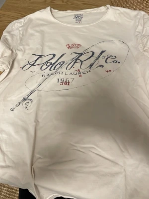 Vit t-shirt från Polo Ralph Lauren - Snygg vit t-shirt från Polo Ralph Lauren med tryck framtill i blått och rött. Klassisk rund halsringning och korta ärmar. Tillverkad i 100% bomull för en skön och luftig känsla. Perfekt till jeans eller shorts för en avslappnad look.