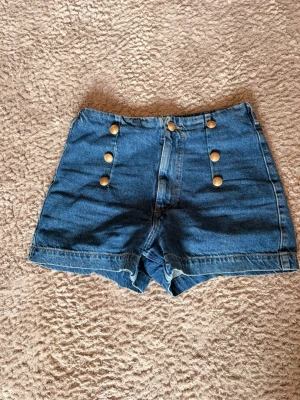 Blå högmidjade jeansshorts med knappar - Snygga blå denimshorts med hög midja och sex guldfärgade knappar framtill. Shortsen har två stora bakfickor och klassisk jeanslook. Perfekta för varma dagar och enkel att matcha med olika toppar.