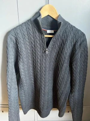 Grå kabelstickad tröja från Esprit - Kabelstickad grå tröja från Esprit med halv dragkedja och hög krage. Tröjan har långa ärmar och ribbade muddar vid ärmslut och nederkant. Perfekt för dig som gillar klassisk och stilren look.