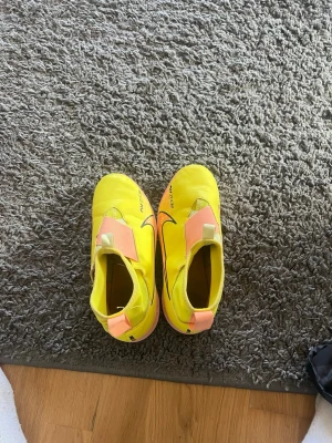 Nike Air Zoom gula fotbollsskor - Säljer ett par Nike Air Zoom fotbollsskor i en riktigt schysst gul färg med rosa detaljer och svart swoosh. Skorna har kardborreband och är tillverkade i syntetmaterial med greppvänlig sula. Perfekta för dig som vill sticka ut på planen!