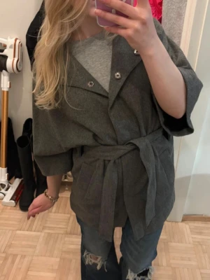 Poncho jacka - Stilren grå rock i oversized modell med bred krage och knytskärp i midjan. Rocken har trekvartsärmar och dubbla knappar framtill. Perfekt för lager-på-lager och en avslappnad look.