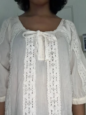 coquette vitt nattlinne  - Söt vit kortklänning i bomull med spetsdetaljer och volanger. Klänningen har trekvartsärm, knytband framtill och vacker broderad spets längs ärmar och framstycke. Perfekt för en romantisk och somrig look.