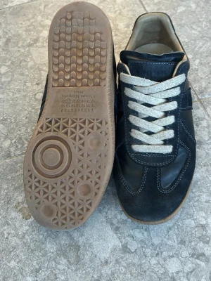 Maison margiela - Snygga svarta sneakers med beige sula och vita snören. Ovandelen är i en mix av mocka och läder, med klassisk retrodesign och kontrastsömmar. Perfekta för dig som gillar stilrena och tidlösa sneakers med en sportig vibe.