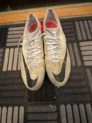 Nike Mercurial fotbollsskor vit/röd - Säljer ett par Nike Mercurial fotbollsskor i vitt med svarta detaljer och röd häl. Skorna har snörning, låg profil och är tillverkade i syntetmaterial för bra grepp och känsla på planen. Perfekta för dig som vill ha snabba och lätta skor.