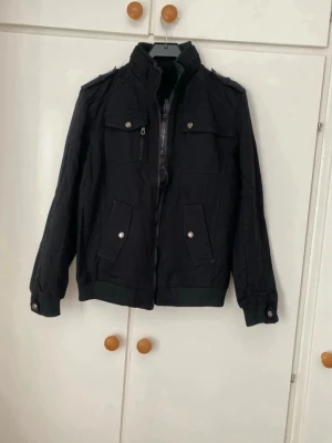 Svart jacka med fickor - Snygg svart jacka med hög krage, axelklaffar och flera fickor med tryckknappar. Jackan har ribbade muddar vid ärmslut och nederkant samt dragkedja framtill. Perfekt för dig som gillar en stilren och enkel look.