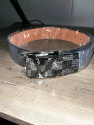 Rutmönstrat Louis Vuitton bälte - Snyggt bälte från Louis Vuitton med klassiskt rutmönster i svart och grått. Bältet har en silverfärgad LV-logga som spänne och insidan är ljusbrun. Perfekt accessoar för att lyfta din outfit.